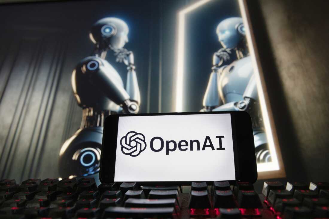 In front is a black keyboard with red backlighting. In the back is a horizontal tablet, displaying the OpenAI name and the tech company's logo on the screen. In the back is a humanoid metal robot looking into a mirror.