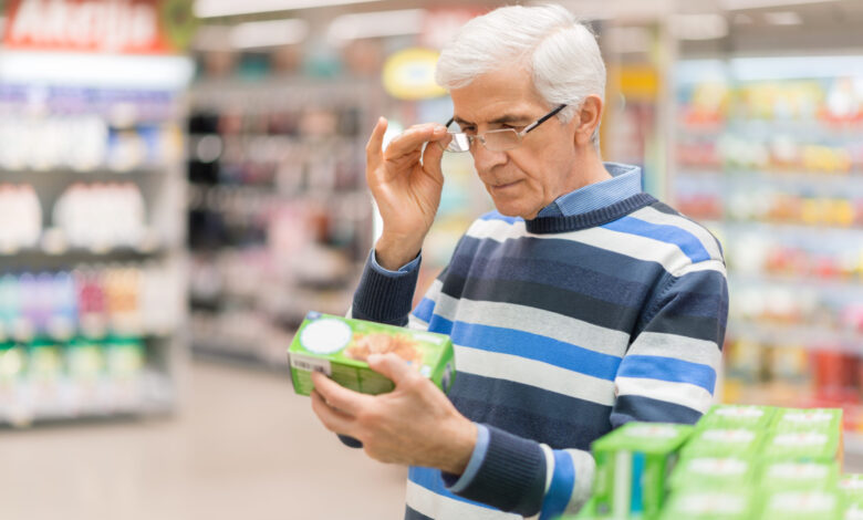 Older man in striped sweater stands in grocery store reading the label on a green box of food