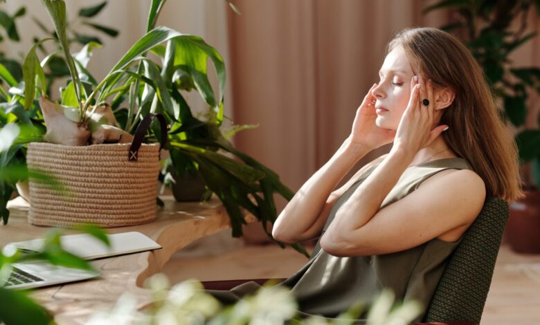 Woman sits in room surrounded by houseplants, eyes closed, rubbing temples