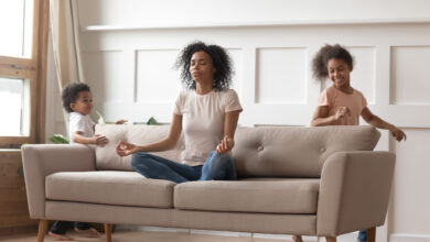 Mom sitting cross-legged on sofa in a meditation pose with two children running around the sofa