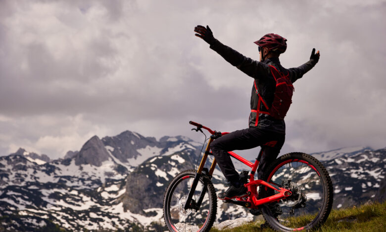 Male mountain biker stands with arms outstretched on mountaintop astride red bike