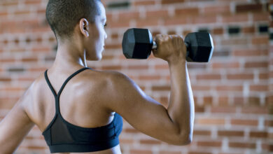 Woman wearing black sports bra is lifting dumbbell while facing a brick wall.