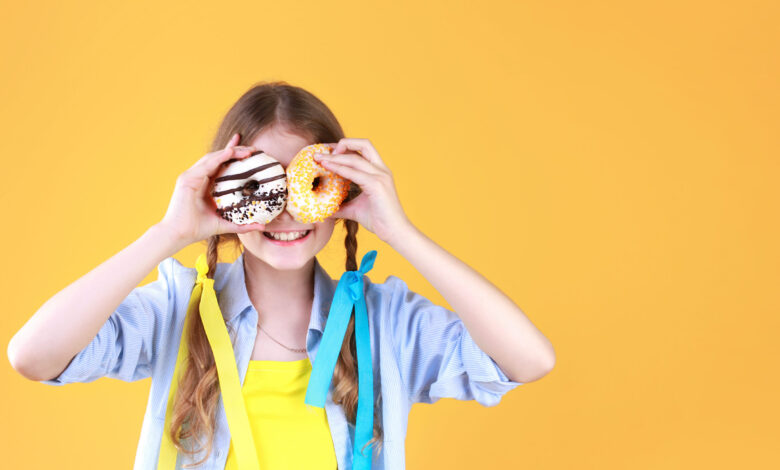 Girl in pigtails standing in front of a yellow background smiling and holding donuts in front of her eyes