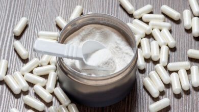 Open container of white powder with scoop on wooden table surrounded by white supplement capsules.
