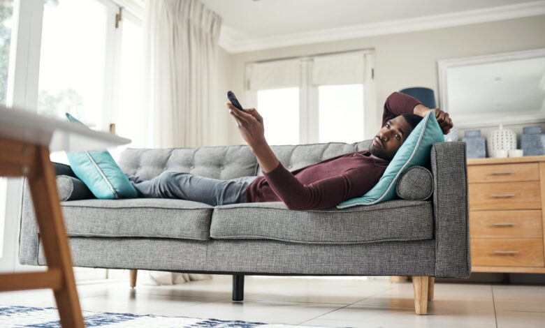 Shot of a man using a remote control while lying on the couch at home.