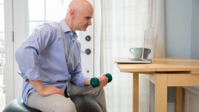 Man sitting on exercise ball holding dumbell in front of laptop.