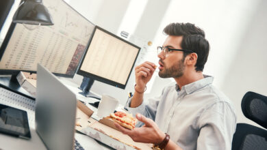 Man sitting at bank of computers eating pizza and looking stressed.