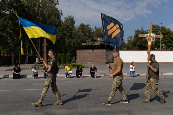 Three soldiers march down a street, with two carrying flags and another carrying a cross during a funeral.